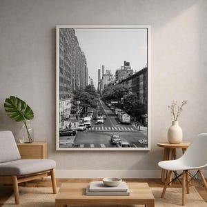 May include: Monochrome photograph of a city street. The image shows a wide avenue with tall buildings, trees, and traffic. A white framed print hangs on a grey wall, with furniture in the foreground.
