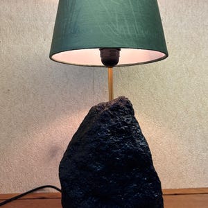 May include: A unique table lamp featuring a dark green lampshade and a black, textured base. The lamp has a gold-colored stem and a black power cord. The lampshade is a deep green color and the base appears to be made of stone.