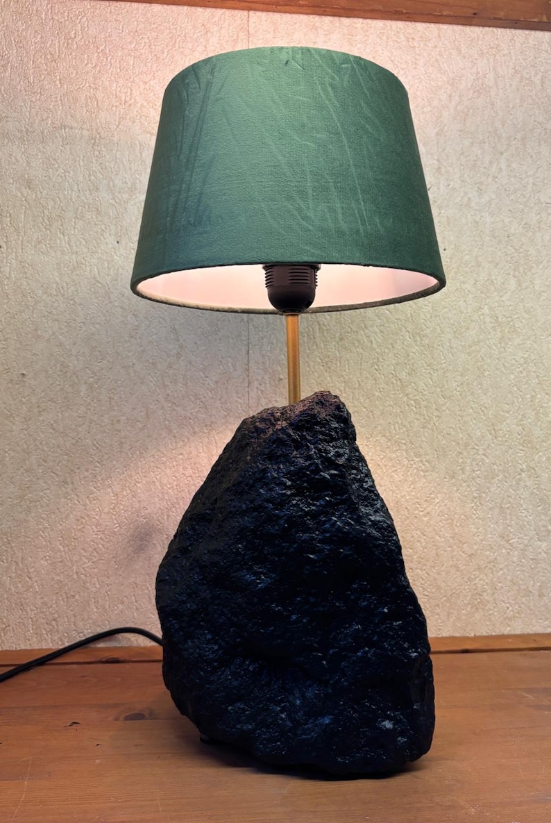 May include: A unique table lamp featuring a dark green lampshade and a black, textured base. The lamp has a gold-colored stem and a black power cord. The lampshade is a deep green color and the base appears to be made of stone.