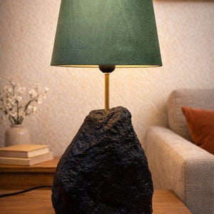 Puede incluir: Lámpara de mesa moderna con pantalla verde oscuro y base negra texturizada. La lámpara tiene un tallo dorado y está sobre una mesa de madera. Un elemento decorativo para el hogar.