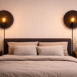 Può includere: Scena di camera da letto con un letto con lenzuola e cuscini dai toni neutri. Due applique circolari, marrone scuro, con lampadine a vista fiancheggiano il letto. Comodini in legno e un vaso decorativo.
