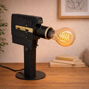 Könnte beinhalten: Eine schwarze und goldfarbene Vintage-Filmkamera-Lampe mit einer großen, leuchtenden Edison-Glühbirne. Die Lampe steht auf einem Holztisch, im Hintergrund eine Pflanze und ein gerahmtes Bild.