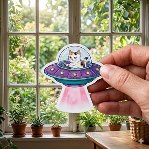 May include: A colorful sticker featuring a calico cat piloting a purple and teal UFO. The UFO has yellow lights and a pink beam. The sticker is held in front of a window with plants.
