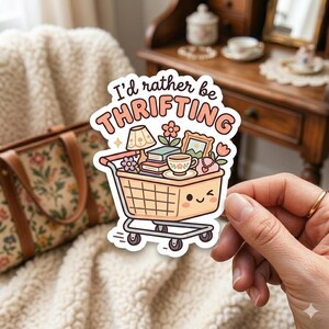 May include: A sticker featuring a shopping cart filled with thrifted items like a lamp, books, and a teacup. The cart has a smiling face and the text "I'd rather be THRIFTING" in a playful font. The sticker is held by a hand.