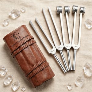 May include: A collection of silver tuning forks with different frequencies, next to a brown leather case with the text "Guided by the Moon" and a crescent moon design. Clear quartz crystals are scattered around the forks and case.