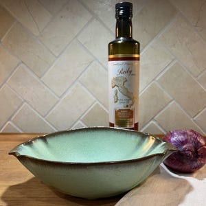 May include: A light green ceramic bowl with a brown rim, a bottle of olive oil, and a red onion. The olive oil bottle has a label with the word "Sicily" and a map of Italy. The bowl is on a wooden surface.