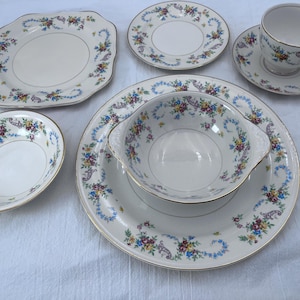 May include: A vintage cream-colored china set with floral and gold trim. The set includes a large plate, a bowl, a small oval dish, two smaller plates, a teacup, and a saucer. The floral design features blue, yellow, and purple flowers.
