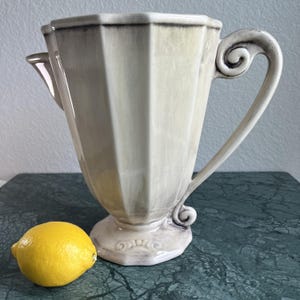 May include: A cream-colored, vintage-style pitcher with a unique, angular design and a decorative handle. The pitcher has a spout and sits on a pedestal base. A bright yellow lemon rests beside the pitcher, adding a pop of color.