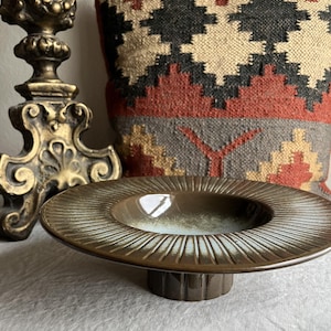 May include: A brown ceramic pedestal bowl with a ridged rim and a light blue interior. The bowl sits on a short, fluted base. A gold-colored decorative object is to the left, and a patterned pillow is in the background.