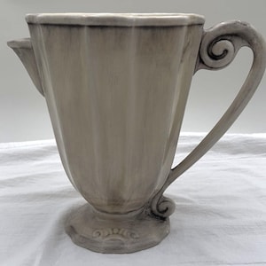 May include: A light gray ceramic pitcher with a decorative handle and spout. The pitcher has a fluted body and a detailed base. The handle features a scroll design. The pitcher is likely used for serving beverages.
