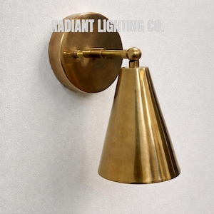 May include: A brass wall sconce featuring a conical shade and a circular backplate. The sconce is mounted on a white wall. The text "RADIANT LIGHTING CO." is printed on the backplate.