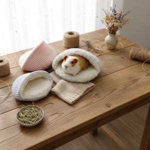 May include: A guinea pig in a white and grey pet bed. The image shows three pet beds in different colours: pink, blue, and white. A small bowl of hay and a beige knitted blanket are also on the wooden table.