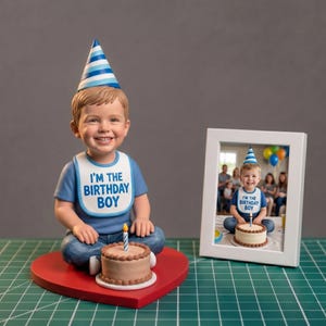 Könnte beinhalten: Figur eines lächelnden Jungen mit blauem Hemd, Lätzchen mit dem Text "I'M THE BIRTHDAY BOY" und einer Partyhut. Er sitzt auf einer herzförmigen roten Basis mit einem kleinen Kuchen. Ein gerahmtes Foto zeigt denselben Jungen.