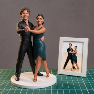 May include: A figurine of a dancing couple in formal attire. The man is in a black shirt and trousers, and the woman wears a teal dress. A framed photo of the couple is on the right. The figurine is on a white circular base.