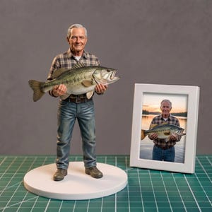 May include: A figurine of an older person holding a large fish, with a framed photo of the same person holding a fish. The figurine is on a white base, and the photo is in a white frame. The person is wearing a plaid shirt and jeans.