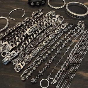 Könnte beinhalten: Eine Auswahl an Silberarmbändern mit verschiedenen Designs, darunter Kettenglieder, Kreuzmotive und verzierte Verschlüsse. Die Armbänder sind auf einer dunklen Holzoberfläche angeordnet, die ihre detaillierte Verarbeitung und Texturen hervorhebt.