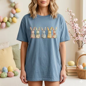 Puede incluir: Una camiseta azul con un diseño de seis ilustraciones de conejos coloridos. Cada conejo tiene un patrón y una pajarita diferentes. La camiseta es de estilo casual y cómodo, perfecta para Semana Santa o primavera.