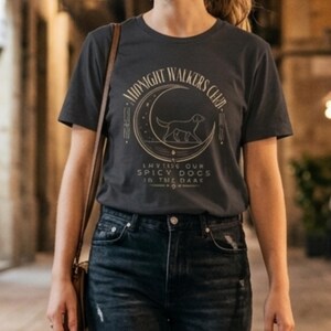 Puede incluir: Una camiseta de manga corta gris carbón con un gráfico dorado de una luna creciente, un perro y el texto "MIDNIGHT WALKERS CLUB" e "INVITING OUR SPICY DOGS IN THE DARK" impreso en la parte frontal del pecho de la prenda.