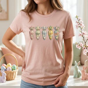 Puede incluir: Camiseta rosa claro con un diseño de fila de zanahorias coloridas. Cada zanahoria tiene un patrón y un lazo únicos. La camiseta es de estilo casual, perfecta para Pascua o primavera. Una cesta de huevos de Pascua y conejitos decorativos en el fondo.