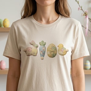 Puede incluir: Una camiseta beige con un diseño de Pascua. El diseño incluye un conejito, una zanahoria, un huevo y un pollito, todos con patrones de tela acolchada suave. Decoraciones de Pascua son visibles en el fondo.