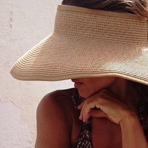 Könnte beinhalten: Ein beigefarbener, gewebter Visor mit breiter Krempe, der Schatten spendet. Der Visor wird von einer Person getragen, die ihr Gesicht teilweise verdeckt. Die Person trägt ein gemustertes Oberteil. Der Hintergrund zeigt eine weiße Wand und Steinstufen.