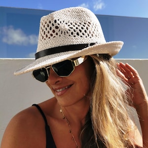 Op de afbeelding: Een witte geweven fedora hoed met een zwarte band en een goudkleurige zonnebril. De hoed heeft een geperforeerd ontwerp. De persoon draagt een zwarte top en een gouden ketting. De achtergrond is een helderblauwe lucht.