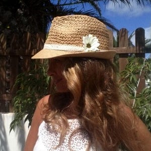 Op de afbeelding: Een strooien fedora hoed met een witte madeliefbloem en een witte kanten band. De hoed heeft een brede rand en een geweven textuur. De hoed is beige en crèmekleurig. Een zomeraccessoire.
