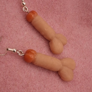May include: A pair of novelty earrings featuring a phallic design. The earrings are flesh-toned with a reddish-orange tip and a silver-colored hook. The earrings are displayed against a pink background.
