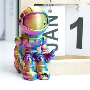 Op de afbeelding: Een kleurrijk astronautenbeeldje met een regenboog metallic afwerking. Het speelgoed staat voor een kalender met de tekst "JAN 1". De astronaut heeft een helm, een lichaam, armen en benen. Het beeldje heeft een levendige kleurenmix.