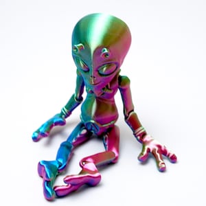 Op de afbeelding: Een kleurrijk, beweeglijk alien-beeldje in tinten blauw, groen, roze en goud. De alien heeft een grote kop en slanke ledematen en zit. Het figuur is gemaakt van een glanzend, iriserend materiaal.