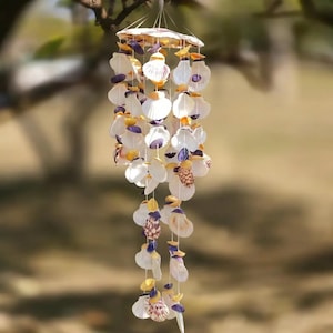 May include: A hanging seashell wind chime. The chime features numerous white, cream, purple, and yellow seashells strung together. The top has a circular design, and the shells hang in multiple strands. The background is blurred, suggesting an outdoor setting.