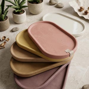 May include: A stack of oval-shaped decorative trays in various colors: pink, yellow, and brown. The trays are made of a solid material and have a smooth surface. The trays are stacked on top of each other. Small potted plants and stones are in the background.