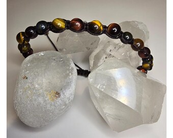 May include: A beaded bracelet featuring tiger's eye stones in shades of brown, gold, and black. The beads are strung on a black cord, creating a circular shape. The bracelet is displayed on a white surface with clear quartz crystals.