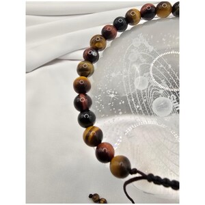 May include: A beaded bracelet featuring tiger's eye stones in shades of brown, red, and black. The bracelet is strung on a black cord and is displayed on a white surface.