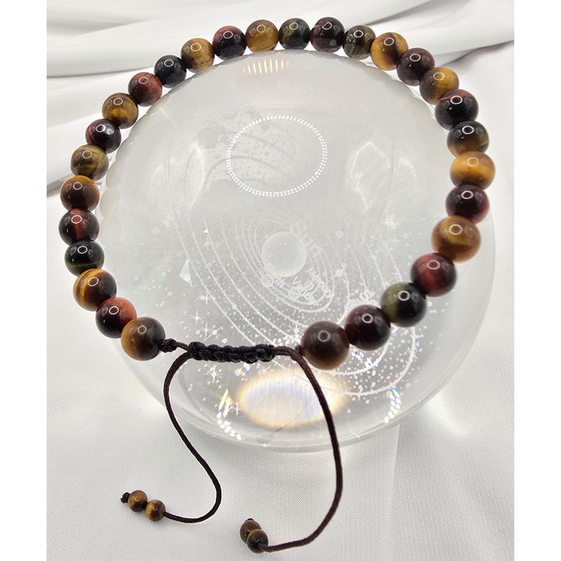 May include: A beaded bracelet featuring tiger's eye gemstones in shades of brown, gold, and red. The bracelet is strung on a black cord with an adjustable sliding knot closure. The gemstones are round and polished, with a natural, earthy appearance.