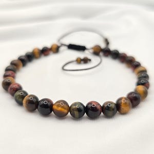 May include: A beaded bracelet featuring tiger eye stones in shades of brown, gold, and red. The bracelet is strung on a black cord with an adjustable sliding knot closure. The beads are approximately 0.3 inches in diameter.