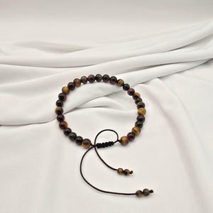 May include: A beaded bracelet featuring a mix of brown, yellow, and red tiger eye stones. The bracelet is strung on a dark brown cord with an adjustable sliding knot closure. The beads are approximately 0.3 inches in diameter.