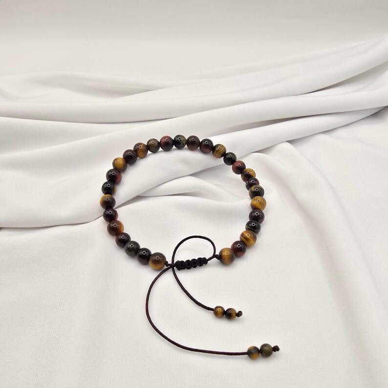 May include: A beaded bracelet featuring a mix of brown, yellow, and red tiger eye stones. The bracelet is strung on a dark brown cord with an adjustable sliding knot closure. The beads are approximately 0.3 inches in diameter.