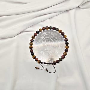 May include: A beaded bracelet featuring tiger's eye stones in shades of brown, red, and gold. The bracelet is strung on a black cord with an adjustable sliding knot closure. The beads are approximately 0.8 cm in diameter.