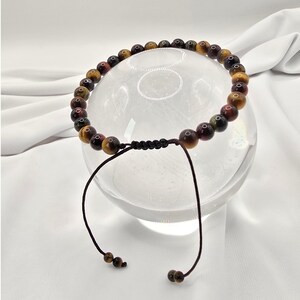 May include: A beaded bracelet featuring tiger's eye stones in shades of brown, gold, and red. The bracelet is strung on a black cord with adjustable brown string ties. The beads are approximately 0.3 inches in diameter.