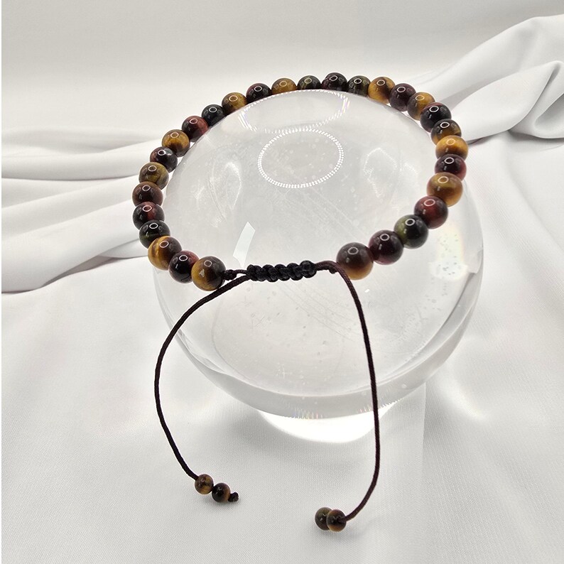 May include: A beaded bracelet featuring tiger's eye stones in shades of brown, gold, and red. The bracelet is strung on a black cord with adjustable brown string ties. The beads are approximately 0.3 inches in diameter.
