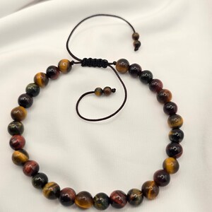May include: Adjustable bracelet featuring a strand of polished, multicolored tiger eye beads. The beads display a mix of brown, yellow, red, and black hues. The bracelet has a black cord with a sliding knot closure for a custom fit.