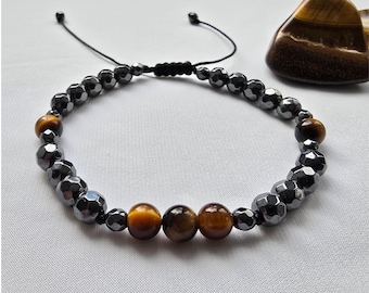 May include: A beaded bracelet featuring faceted, dark gray hematite beads and round, golden tiger's eye beads. The bracelet has an adjustable black cord closure. A tiger's eye stone is in the background.