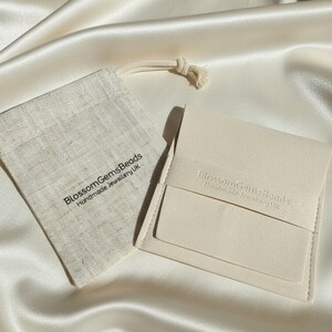May include: A close-up shot of a cream-colored jewelry pouch and a small drawstring bag. The pouch is rectangular with a flap closure. The bag and pouch have the text "BlossomGemsBeads Handmade Jewellery UK". Both items are on a satin surface.