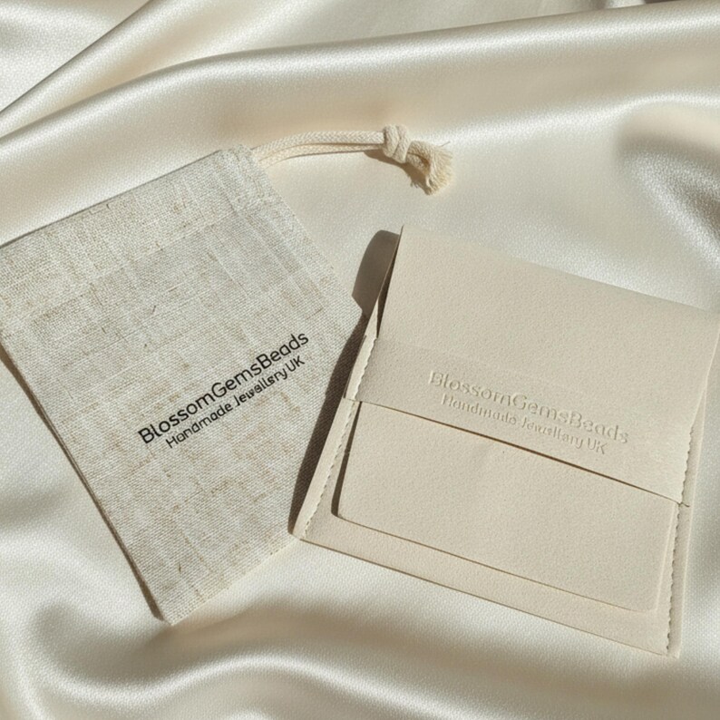May include: A close-up shot of a cream-colored jewelry pouch and a small drawstring bag. The pouch is rectangular with a flap closure. The bag and pouch have the text "BlossomGemsBeads Handmade Jewellery UK". Both items are on a satin surface.