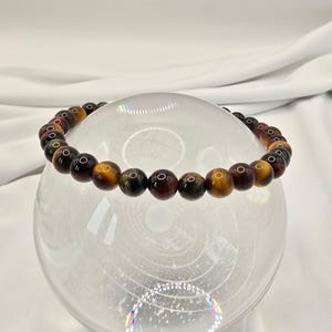 May include: A beaded bracelet featuring polished tiger eye stones in shades of brown, gold, and burgundy. The bracelet is strung on an elastic band and is displayed on a clear glass sphere. The stones are approximately 0.3 inches in diameter.
