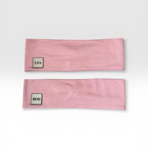 May include: Two light pink headbands. Each headband has a small, square, silver-colored tag with initials. One tag reads "CDA" and the other reads "RDH". The headbands are made of a soft, textured fabric.