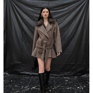 May include: A brown wool coat featuring a faux fur collar and a belted waist. The coat has a double-breasted design and a ruffled hem. The model is wearing black knee-high boots. The background is a black backdrop.