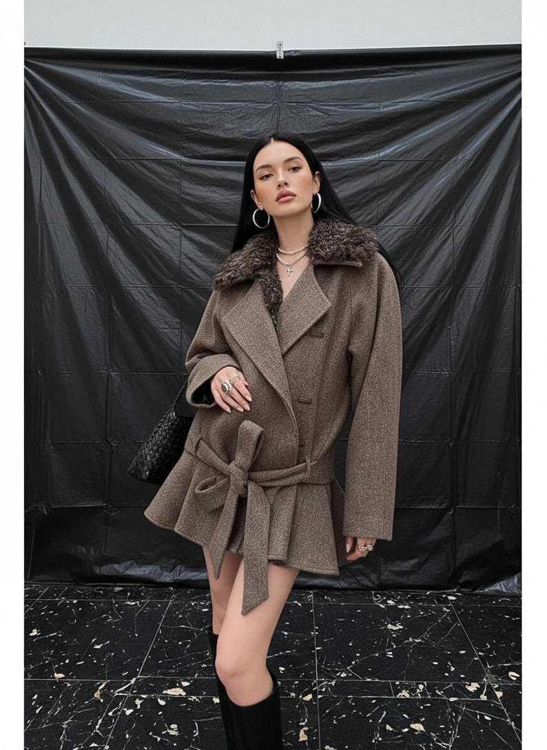 May include: A brown coat with a faux fur collar and a belted waist. The coat has a double-breasted design and a flared skirt. The model is wearing black boots and hoop earrings. The background is a black textured surface.