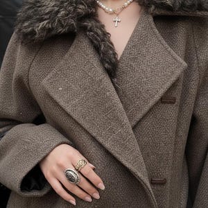 May include: A brown wool coat featuring a faux fur collar. The coat has a double-breasted design with large lapels and two brown buttons. A pearl necklace with a cross pendant and multiple rings are also visible.